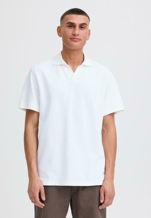 Jonge man met kort haar, gekleed in een effen wit poloshirt met korte mouwen en een donkere broek, staand tegen een lichte achtergrond.