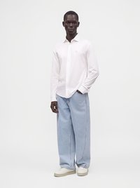 Homme debout portant une chemise blanche à manches longues avec boutons, un jean large bleu clair et des baskets blanches sur un fond uni.