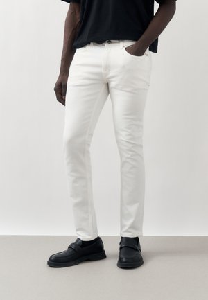 Mand iført slim-fit hvide jeans, sorte loafers og en sort t-shirt, står med den ene hånd i lommen mod en ensfarvet baggrund.