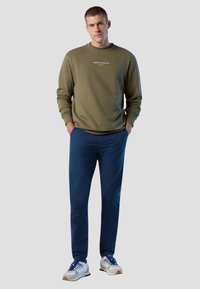 Felpa khaki con logo bianco, pantaloni blu con una superficie liscia e sneakers bianche con accenti blu. Vestibilità complessiva rilassata.