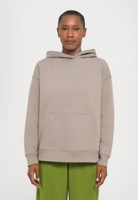 Sweat-shirt à capuche en taupe clair, avec une poche kangourou, des poignets côtelés et une coupe décontractée. Porté avec un pantalon vert. Texture lisse.