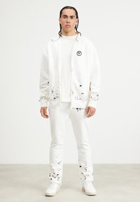 Hvit zip-up hettegenser med fargeplask og et emblem. Sammen med matchende hvite bukser med plask. Glatt tekstur, avslappet passform.