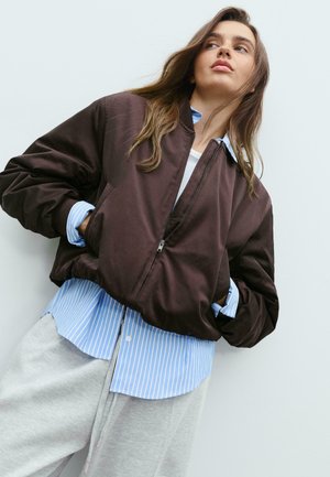 Giacca bomber marrone con zip frontale, polsini a coste e due tasche, indossata sopra una camicia a righe blu chiaro con bottoni e pantaloni della tuta grigi.