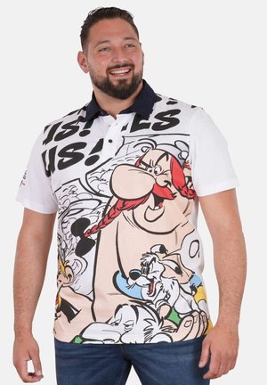 Homme souriant portant un polo blanc avec un grand dessin coloré de personnage de dessin animé et un col sombre, assorti à un jean bleu.