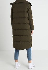 Olivenfarvet pufferjakke, knælang, vandret quiltet mønster, stor krave, sidelufter, båret over blå jeans og hvide sneakers.