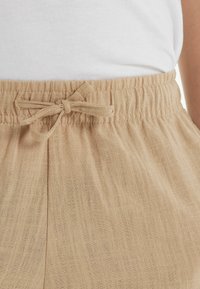 Světlé béžové, texturované tkaniny s elastickým pasem a zavazovací šňůrkou. Obsahuje viditelné švy a měkký, uvolněný tvar.