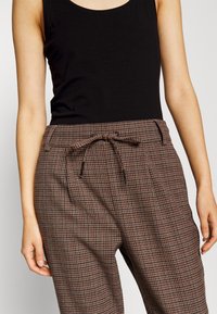 Hnědé kostkované kalhoty s nastavitelným pasem, bočními kapsami a volným střihem, vyrobené z lehkého materiálu s hladkou texturou.