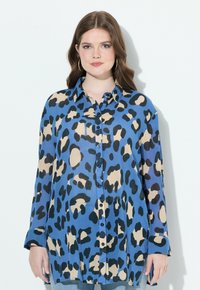 Giovane donna con pelle chiara e capelli castani ondulati che indossa una camicia a maniche lunghe con stampa leopardata blu, beige e nera e orecchini a cerchio.