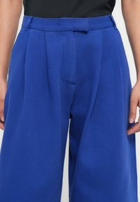 Blaue, weit geschnittene Hosen aus strukturiertem Stoff, mit hoher Taille, Plisseefalten und Gürtelschlaufen. Abgebildet vom Taillenbereich abwärts, mit seitlich hängenden Händen.