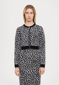 Cardigan à imprimé léopard en noir et blanc, design à fermeture éclair, ourlet et poignets côtelés noirs, silhouette ajustée, texture douce.