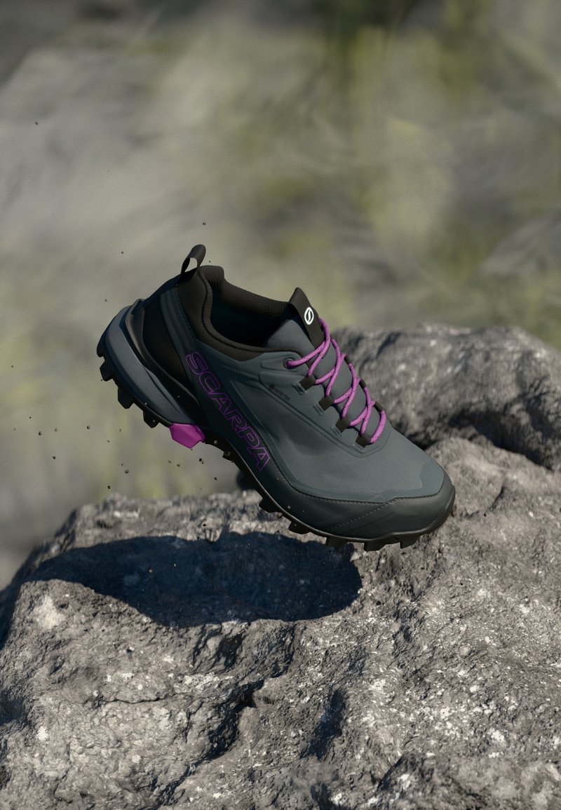 Shoe de caminhada cinza e preto com atacadores roxos e nome da marca, pairando acima de uma superfície rochosa num ambiente ao ar livre.