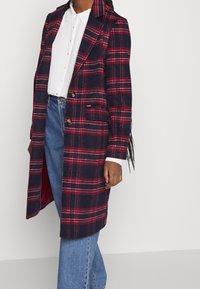 Nainen, jolla on päällään tummansininen ja punainen ruudullinen pitkä takki valkoisen kauluspaita ja sinisten farkkujen päällä, seisoo tasaisen vaaleaa taustaa vasten.