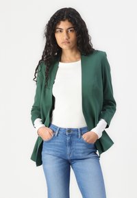 Zelený blazer s tvarovaným designem, zapínání na jeden knoflík a dvě postranní kapsy. Nositý přes bílé tričko a kombinovaný s modrými džíny.