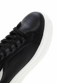 Zapatilla de cuero negra con punta redonda, suela de goma blanca y cordones planos negros. Presenta un diseño distintivo en el panel lateral.