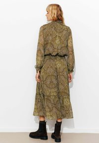 Robe longue à motifs en vert olive avec des designs paisley, dotée de manches ballon, d'une taille élastique et d'un ourlet à volants. Portée avec des bottines noires.
