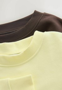 Brune og gule sweatshirts stablet, med ribstrikkede halsudskæringer. Den brune har en blød tekstur, mens den gule er glat og let dæmpet.