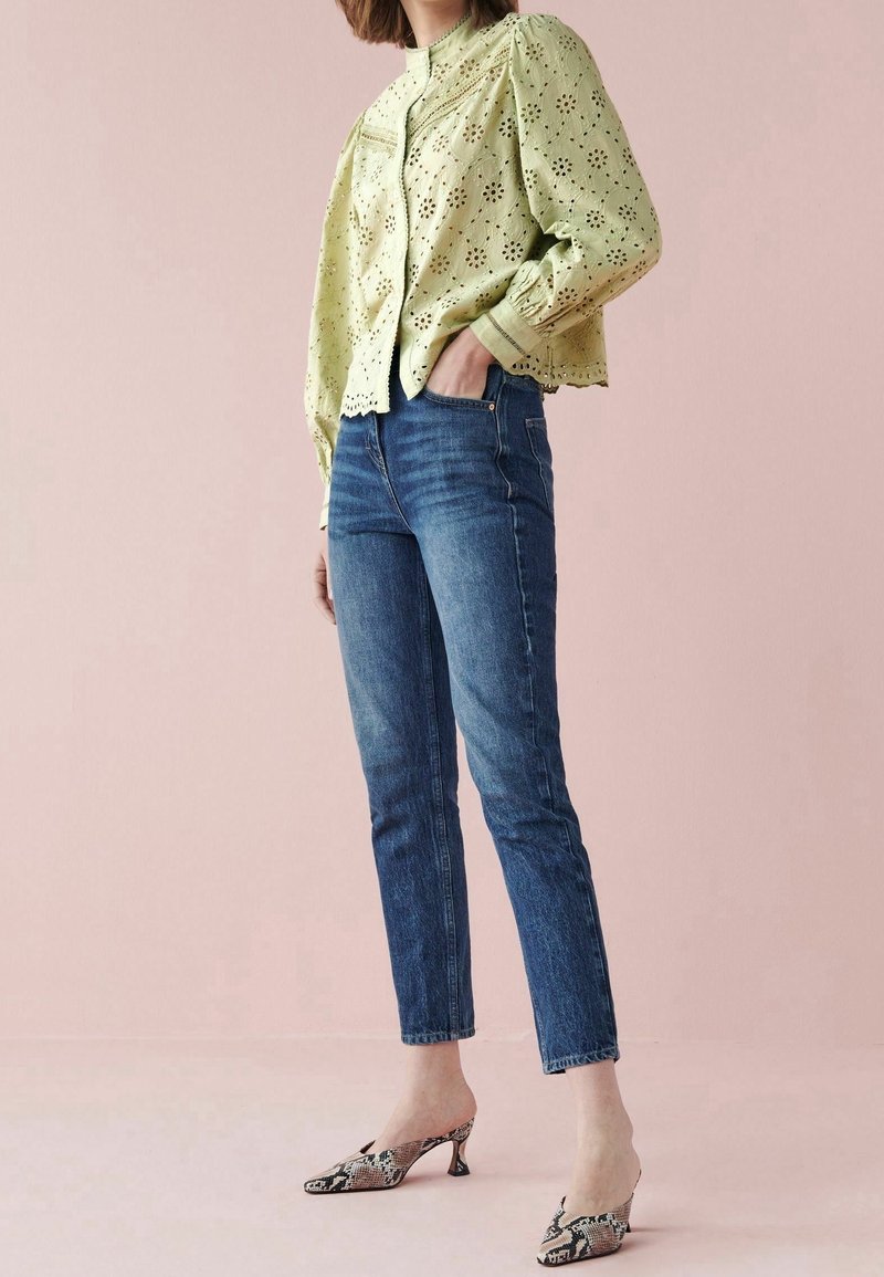 Femme portant un chemisier vert clair ajouré, un jean slim bleu et des escarpins slingback en peau de serpent, debout sur un fond rose pâle.