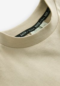 Beige bomull t-shirt med rund halsringning. Etiketten säger "BAKER by Ted Baker" på en mörk bakgrund. Slät textur synlig.