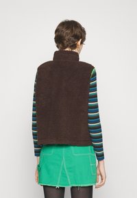 Brauner Fleece-Weste mit hohem Kragen, getragen über einem gestreiften Langarmshirt. Der Rock ist aus grünem Denim mit ausgefranstem Saum und aufgesetzten Taschen auf der Rückseite.