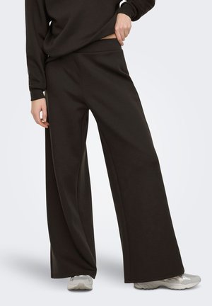 Osoba ubrana w czarne szerokie spodnie, czarną długą bluzkę i szare trampki, z jedną ręką poprawiającą pas spodni.