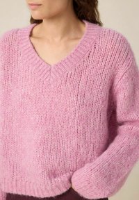 Pull en maille épaisse rose avec col en V, featuring un fil texturé et des poignets côtelés. Finition douce et légèrement pelucheuse.