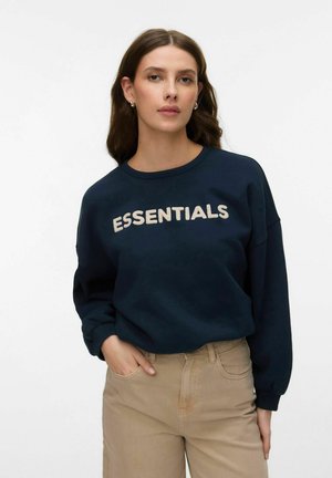 Kvinne iført marineblå genser med teksten "ESSENTIALS" og beige bukser, stående med en hånd i lommen mot hvit bakgrunn.