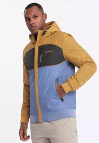 Färgglad jacka med huva i gult, svart bröstpanel och blå nederdel. Har dragkedjefickor och ribbade ärmslut.