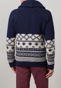 Marinblå stickad tröja med bred krage, prydd med vita geometriska mönster och texturerad tyg. Ribbad muddar och nederkant.
