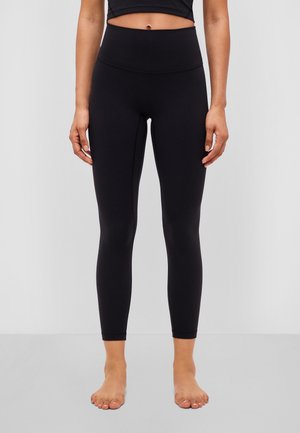 Mujer con leggings negros de cintura alta, de pie descalza frente a un fondo claro y liso.