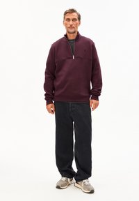 Bordeauxkleurige rits hoodie met een hoge kraag, gestructureerde stof, donkere spijkerbroek en lichtgrijze sneakers. Eenvoudig, casual ontwerp.