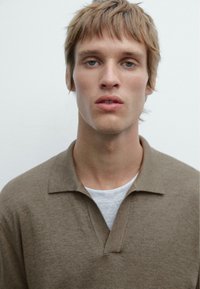 Hnědý pletený polo s límečkem, vrstvený přes světlé šedé tričko. Materiál vypadá měkký, s jemnou texturou.