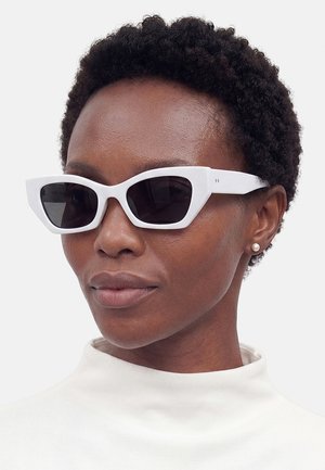 Kobieta nosząca białe okulary przeciwsłoneczne typu cat-eye, perłowe kolczyki i biały top z wysokim kołnierzem, patrząca lekko w bok.
