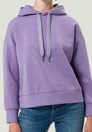 Persona que lleva una sudadera con capucha de color lila claro con cordones anchos y vaqueros azules, mostrada desde el cuello hasta las caderas sobre un fondo liso.