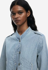 Pavlena košulja s plavim i bijelim prugama, s kragnom, dva prednja džepa i vidljivim detaljima šivanja. Izrađena od lagane tkanine.