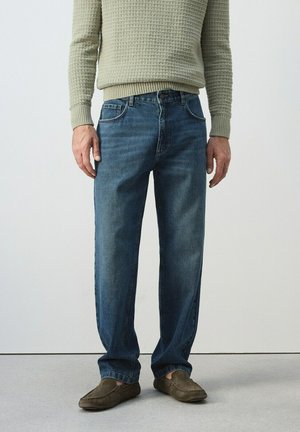 Uomo che indossa un maglione verde chiaro a trama, jeans di velluto a coste blu e scarpe marroni senza lacci, in piedi contro un muro semplice.
