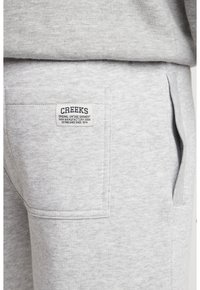 Pantalones de chándal grises hechos de una suave mezcla de algodón, con un bolsillo trasero que tiene una etiqueta que dice "CREEKS" y detalles de costura en tono claro.