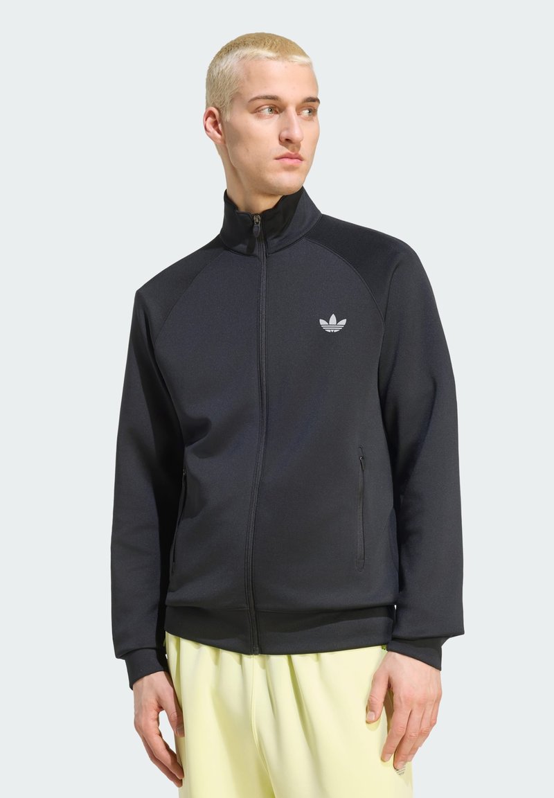 Junger Mann mit kurzen blonden Haaren, der eine schwarze Jacke mit Reißverschluss und Adidas-Logo sowie hellgelbe Hosen trägt, steht vor einem schlichten Hintergrund.