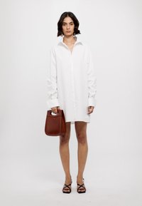 Vit skjortklänning med långa ärmar, oversized passform och breda manschetter. Modellen håller en brun läderhandväska. Svarta sandaler med remmar.