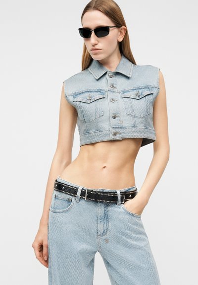 Νεαρή γυναίκα που φοράει στενά μαύρα γυαλιά ηλίου, αμάνικο cropped τζιν μπουφάν, φαρδιά ανοιχτό μπλε τζιν με τρουκς και μαύρη ζώνη με τρουκς.