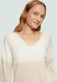 Pull en maille crème à col en V avec une rayure horizontale beige. Il possède des manches longues et un tissu texturé pour un détail supplémentaire.