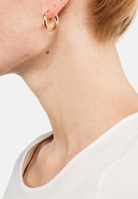 Ohrringe aus roségoldenen Creolen mit polierter Oberfläche, runde Form, mittlere Größe, getragen auf einem hellen Hautton, ergänzt durch einen weißen Ausschnitt.
