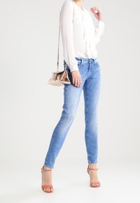 Blusa bianca leggera con volant, abbinata a jeans fitted in denim azzurro. Il modello indossa sandali con tacco alto e cinturino alla caviglia color nude e una borsa metallica.