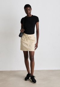 Svart, kortermet, tettsittende t-skjorte kombinert med en beige miniskjørt med snor i livet, brukt med svarte joggesko. Enkel, moderne design.