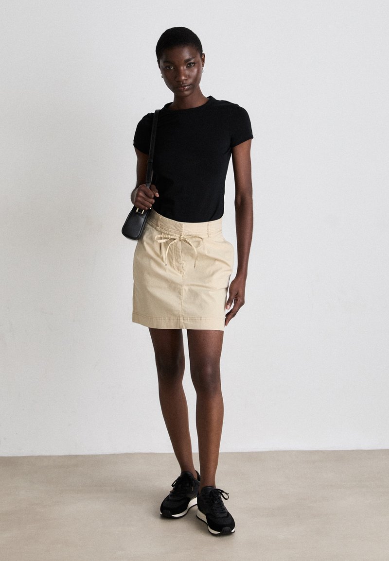 Camiseta ajustada negra de manga corta combinada con una minifalda beige con cintura de cordón, usada con zapatillas negras. Diseño simple y contemporáneo.