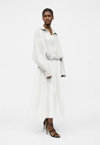 Γυναίκα που φοράει ανοιχτό γκρι φαρδιά ζακέτα, λευκή αέρινη midi φούστα και μαύρα ψηλοτάκουνα σανδάλια, στέκεται μπροστά σε μονόχρωμο φόντο.