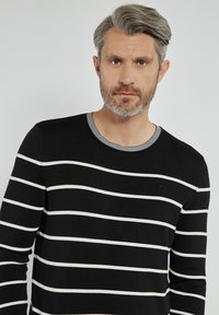 Czarny sweter z długim rękawem i okrągłym dekoltem, z białymi poziomymi paskami oraz szarym prążkowanym kołnierzem. Posiada małe okrągłe logo na klatce piersiowej.