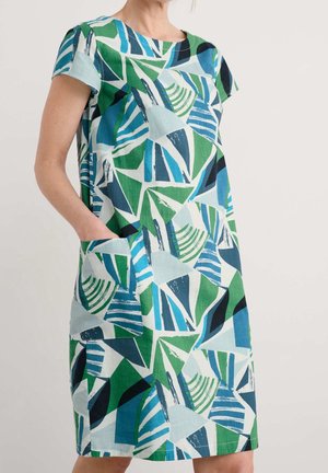 Donna che indossa un vestito corto a maniche corte, lungo fino al ginocchio, con motivo geometrico astratto verde, blu, nero e bianco e tasca frontale.