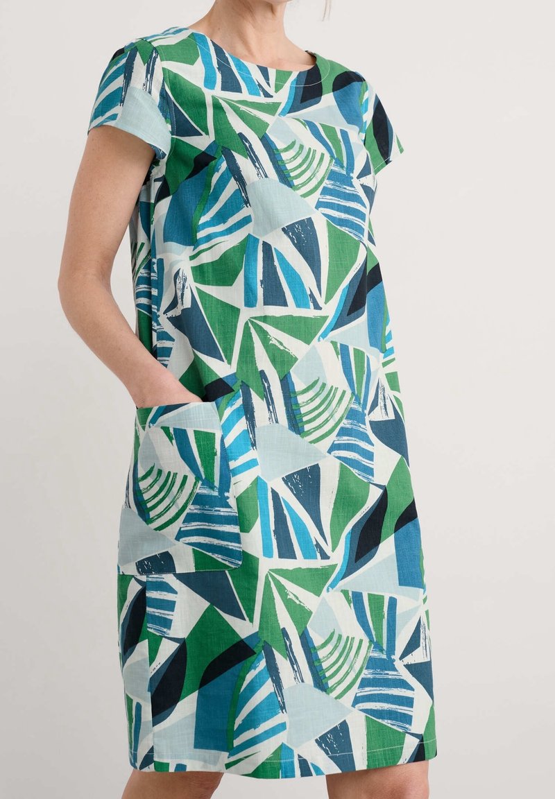 Femme portant une robe courte à manches courtes, longueur genou, avec un motif géométrique abstrait en vert, bleu, noir et blanc et une poche avant.