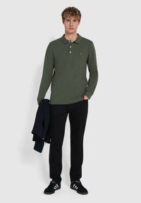 Langarm polos Hemd in grün mit Knopfleiste, getragen mit schwarzen Hosen und einem navyblauen Pullover, der über dem Arm hängt. Es hat weiße Knöpfe und ein Logo.