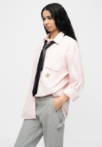 Νεαρή γυναίκα με μακριά μαύρα μαλλιά, που φοράει ένα ανοιχτό ροζ φαρδύ πουκάμισο, μαύρη δερμάτινη γραβάτα και ριγέ παντελόνι, με το χέρι στην τσέπη.